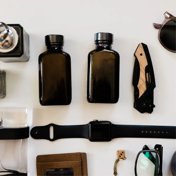 Flach gelegtes EDC-Setup mit brauner Geldbörse, Smartwatch, Taschenmesser mit Holzgriff, Sonnenbrille, dunklen Fläschchen und Accessoires auf weissem Hintergrund.